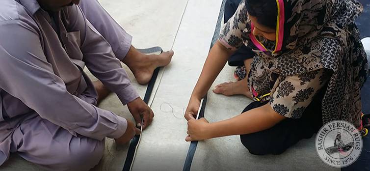 Pakistani rug artisans binding leather strips onto the edges of a carpet to reinforce it.